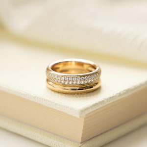 May include: A gold ring with a double band of small, clear gemstones rests on a light-coloured book. The ring has a polished, shiny finish. The background is softly blurred, with a hint of white fabric.