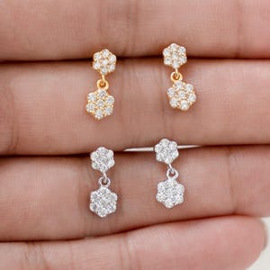 May include: A close-up of two pairs of flower-shaped dangle earrings. One pair is gold-toned, the other is silver-toned. Each earring features a small flower at the top and a larger flower dangling below, both adorned with small, clear gemstones.