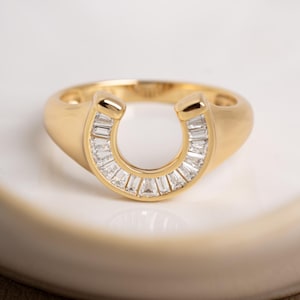 Può includere: Un anello dorato con un design a ferro di cavallo. Il ferro di cavallo è incastonato con piccole pietre chiare rettangolari. L'anello è posto su una superficie bianca. Gioielli.