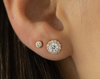 May include: A pair of gold earrings with a small round diamond stud and a larger round diamond with a halo of smaller diamonds.