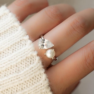 May include: A silver ring featuring a pear-shaped clear gemstone and a white pearl. The ring is worn on a finger.
