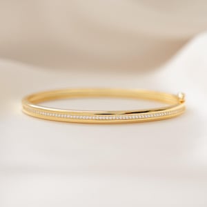 May include: A gold bangle bracelet with a row of small, sparkling stones set into the band. The bracelet is displayed on a white, silky surface, creating a contrast with the warm gold color. The jewelry piece is a classic design.