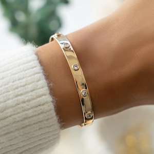 May include: A gold-colored bangle bracelet with clear, round gemstones evenly spaced along the top. The bracelet is worn on a wrist, and the background is blurred, with a white sweater sleeve visible.