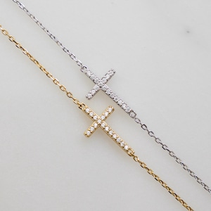 May include: Two delicate cross bracelets, one gold-toned and one silver-toned, each adorned with sparkling gemstones. The crosses are set horizontally on fine chain links, creating a minimalist and elegant design.