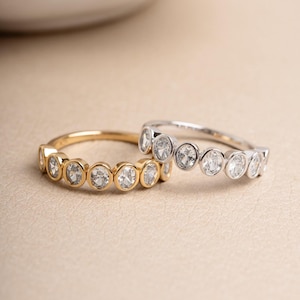 May include: Two rings with clear gemstones. One is gold-toned, the other silver-toned. Each ring has a series of round, clear gemstones set closely together. The rings are displayed on a neutral surface, with a blurred background.