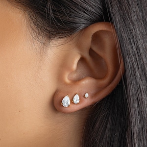 May include: Close-up of an ear with three gold earrings. The earrings are set with clear, pear-shaped gemstones. The largest earring is in the first piercing, followed by a smaller pear-shaped earring, and a tiny round earring.