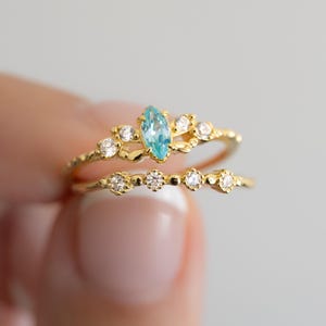 May include: A gold ring with a marquise-shaped blue gemstone surrounded by smaller clear gemstones. The ring is set on a delicate band with a row of smaller clear gemstones.