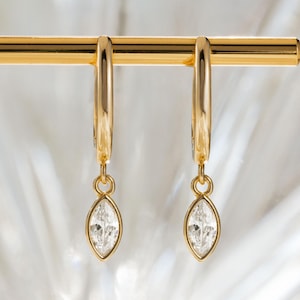 May include: A pair of gold hoop earrings with a marquise-cut, clear gemstone pendant. The earrings are displayed on a gold bar against a blurred, white background. The earrings are a classic jewelry design.