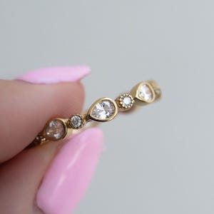 Puede incluir: Un anillo de oro con piedras preciosas claras en forma de pera y redondas alternas. El anillo lo sostiene una persona con esmalte de uñas rosa. Una delicada pieza de joyería.