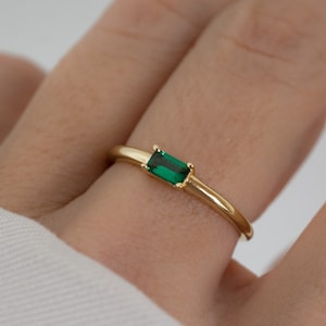 May include: A gold ring with a rectangular emerald-green gemstone. The ring is on a finger, with a white sleeve in the background. The gemstone is set horizontally in the center of the ring.