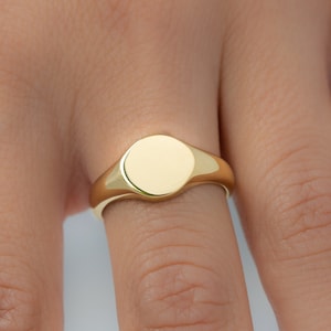 May include: A gold signet ring with a smooth, circular face. The ring is a bright, polished gold color and is worn on a finger. The ring's design is simple and classic, suitable for everyday wear. The ring is a piece of jewelry.