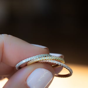 Pode incluir: Três finas alianças de eternidade de diamantes empilhadas em um dedo. As alianças são de diferentes cores de metal: prata, ouro rosa e ouro amarelo.