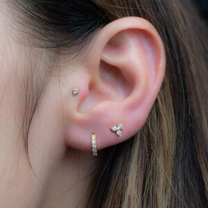 May include: A person's ear with three different earrings. The top earring is a small stud, the middle earring is a small cluster of three stones, and the bottom earring is a small hoop with stones.