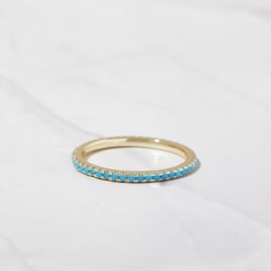 Puede incluir: Un fino anillo de banda de oro con una fila de piedras azul turquesa alrededor de toda la banda.