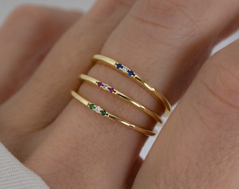 May include: Three gold-colored rings stacked on a finger. Each ring has a row of small, colorful gemstones: blue, red, and green. The rings are thin and delicate, with a polished finish. The background is a soft, neutral color.