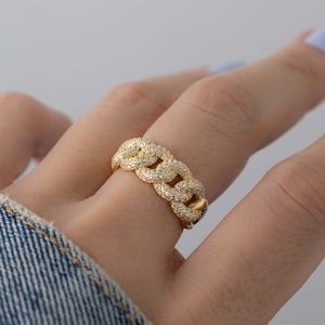 Puede incluir: Un anillo dorado con un diseño de eslabones de cadena, incrustado con pequeñas piedras preciosas transparentes. El anillo se lleva en un dedo, mostrando sus intrincados detalles y su apariencia brillante. El diseño de bucle continuo del anillo crea un aspecto audaz y elegante.