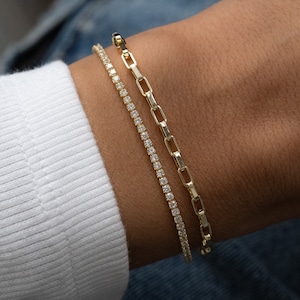 May include: Two gold bracelets on a wrist. One bracelet is a delicate chain with small diamonds, the other is a thicker chain with larger links.
