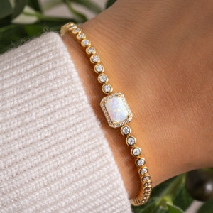May include: A gold bracelet with a white opal gemstone surrounded by small clear stones. The bracelet is on a person's wrist.