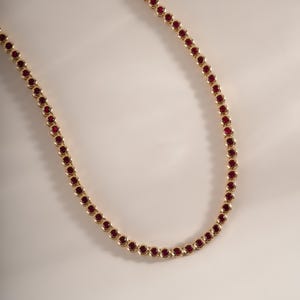 May include: A gold-toned tennis necklace featuring a continuous row of round, deep red stones set in individual metal bezels. The necklace is arranged in a gentle curve, showcasing a uniform design of linked circular settings with vibrant red gems.