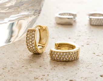 May include: Gold-tone hoop earrings encrusted with small, clear crystals. The earrings are displayed on a light-colored, textured surface, with a blurred glass object in the background. Two additional silver-colored earrings are also visible.