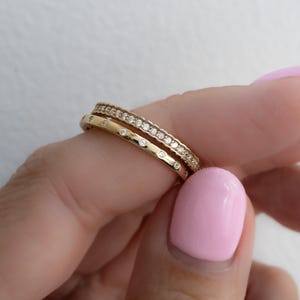 Puede incluir: Dos anillos de oro con pequeñas piedras preciosas transparentes. Un anillo tiene una fila de piedras preciosas, y el otro tiene piedras preciosas espaciadas uniformemente. Los anillos son sostenidos por una persona con esmalte de uñas rosa.