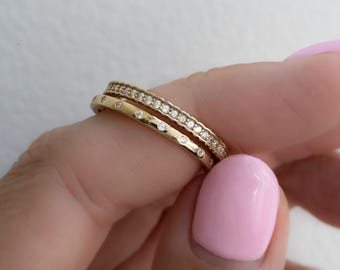 Könnte beinhalten: Zwei goldene Ringe mit kleinen, klaren Edelsteinen. Ein Ring hat eine Reihe von Edelsteinen, der andere hat gleichmäßig verteilte Edelsteine. Die Ringe werden von einer Person mit rosa Nagellack gehalten.