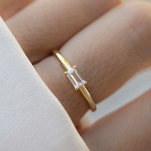 Peut inclure: Une fine bague en or ornée d'une pierre précieuse rectangulaire et transparente. La bague a un anneau fin et poli, et la pierre est sertie dans un style minimaliste. La bague est présentée sur un doigt, sur un fond doux et neutre.