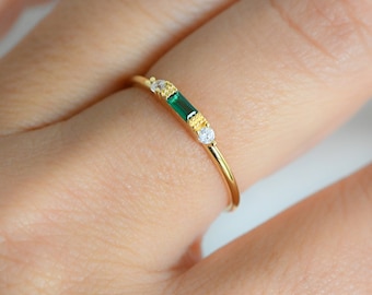 May include: A delicate gold ring featuring a rectangular emerald-cut green gemstone, flanked by two smaller white gemstones. The ring's band is thin and polished, creating a minimalist and elegant design.