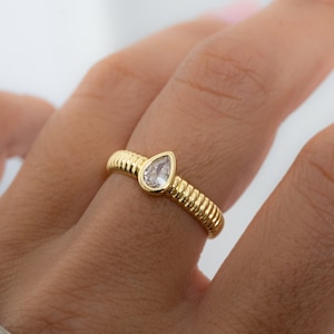 May include: A gold ring with a pear-shaped diamond set in the center. The band is textured with a ribbed design.