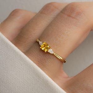 Op de afbeelding: Een gouden ring met een vierkante, gele edelsteen en twee peervormige, witte edelstenen. De ring heeft een gedraaide band. De ring wordt op een vinger getoond.