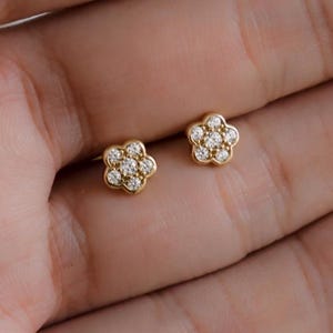 May include: A pair of small, gold-colored flower-shaped stud earrings. Each earring features a cluster of small, clear gemstones at the center, creating a sparkling effect. The earrings are displayed on a light-toned background.