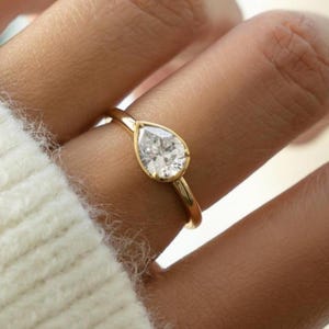 Puede incluir: Un anillo de oro con un diamante en forma de pera. El anillo tiene una delicada banda de oro. El diamante es claro y brillante. El anillo se muestra en un dedo, con un suéter blanco en el fondo.