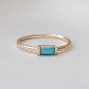 May include: A delicate gold ring featuring a rectangular turquoise stone at the center. Small, clear stones are set on either side of the turquoise. The ring has a thin band and a minimalist design, perfect for everyday wear.
