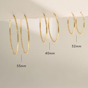 Puede incluir: Pendientes de aro dorados en tres tamaños: 53 mm, 40 mm y 32 mm. Los pendientes tienen una superficie texturizada y se muestran sobre un fondo neutro. Son una joya.