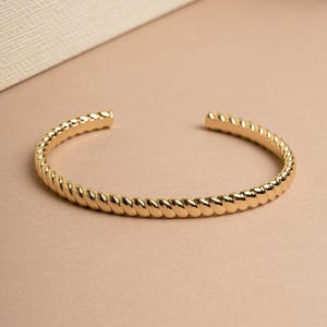 Op de afbeelding: Een goudkleurige armband met een gedraaid touwontwerp. De open manchetarmband is gemaakt van een glanzend, metallic materiaal. De armband is een eenvoudig, elegant sieraad.