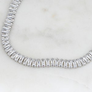 Pu&ograve; includere: Una collana d'argento con una fila di cristalli trasparenti a forma di baguette.