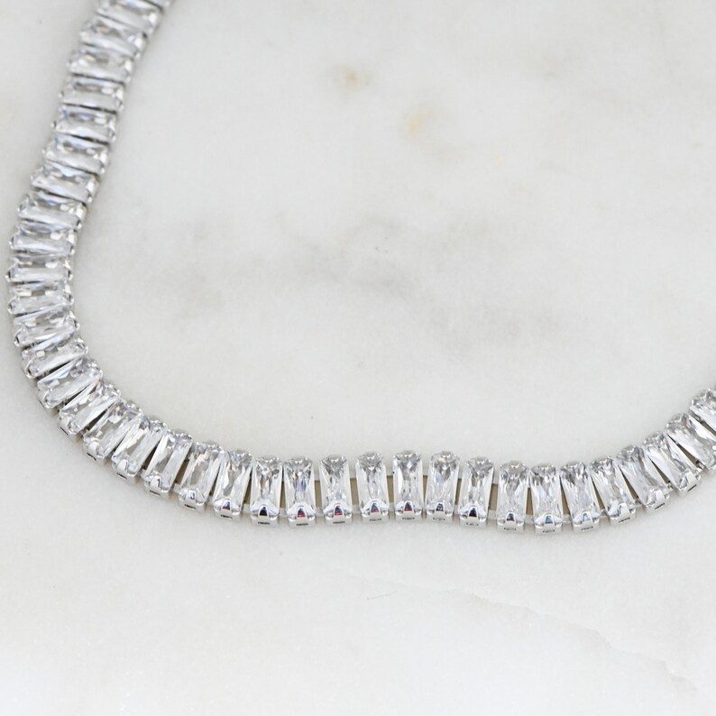 Pu&ograve; includere: Una collana d'argento con una fila di cristalli trasparenti a forma di baguette.