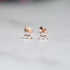 May include: A pair of rose gold stud earrings with three small, clear cubic zirconia stones set in a triangular shape.
