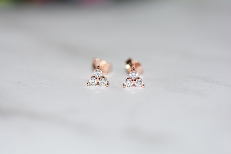May include: A pair of rose gold stud earrings with three small, clear cubic zirconia stones set in a triangular shape.