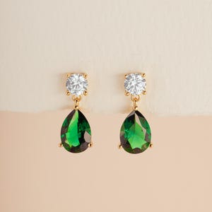 May include: A pair of gold-tone drop earrings featuring a round, clear, faceted stone at the top and a larger, pear-shaped, deep green faceted stone suspended below. The stones are held in place by gold-tone metal prongs and settings.