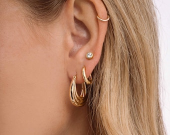 May include: Close-up of a person's ear adorned with gold jewelry. The ear features a small stud earring with a clear stone, a small hoop earring, and a set of three graduated hoop earrings. The earrings are all gold-toned and create a layered effect.