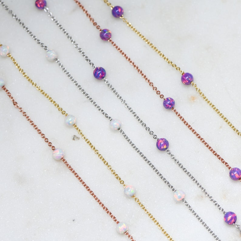 May include: Five delicate necklaces with alternating opal and iridescent purple beads. The necklaces are layered on a white surface.