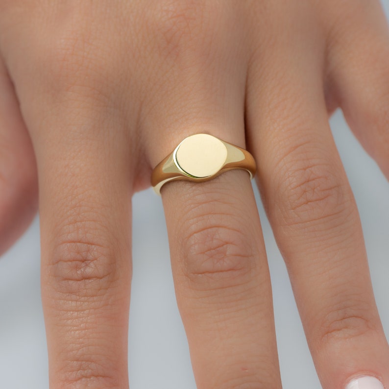 May include: A gold signet ring with a smooth, circular face. The ring is a bright, polished gold color and is worn on a finger. The ring's design is simple and classic, suitable for everyday wear. The ring is a piece of jewelry.