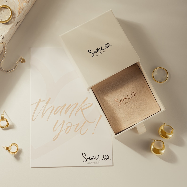 Puede incluir: Una caja blanca con un caj&oacute;n que dice "Sami Jewels" en la parte delantera y en el interior. El caj&oacute;n est&aacute; abierto y tiene un interior beige con el mismo logotipo. Una tarjeta de agradecimiento con el mismo logotipo est&aacute; sobre una superficie blanca. Hay pendientes y anillos dorados sobre la superficie.