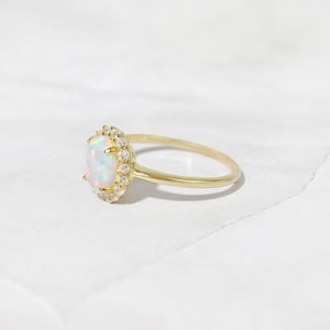 Pode incluir: Um anel de ouro com uma gema de opala branca cercada por um halo de gemas menores e transparentes.