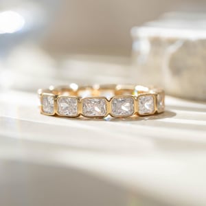 May include: A gold band ring featuring a series of square-cut, clear gemstones. The ring is set in a gold band, with each gemstone individually set. The ring is displayed on a light-colored surface, with soft lighting.