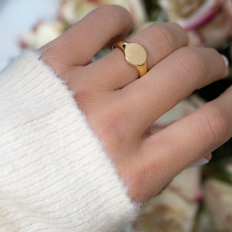 May include: A gold signet ring with a smooth, oval face. The ring is worn on a finger, and the gold reflects light. The ring is a classic piece of jewelry.
