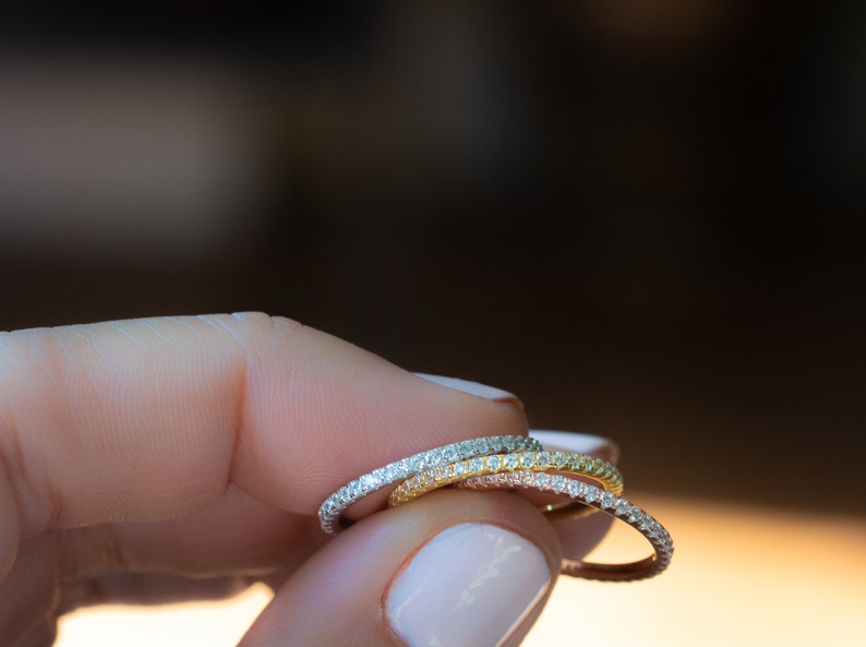 May include: Three thin diamond eternity bands stacked on a finger. The bands are in different metal colors: silver, rose gold, and yellow gold.