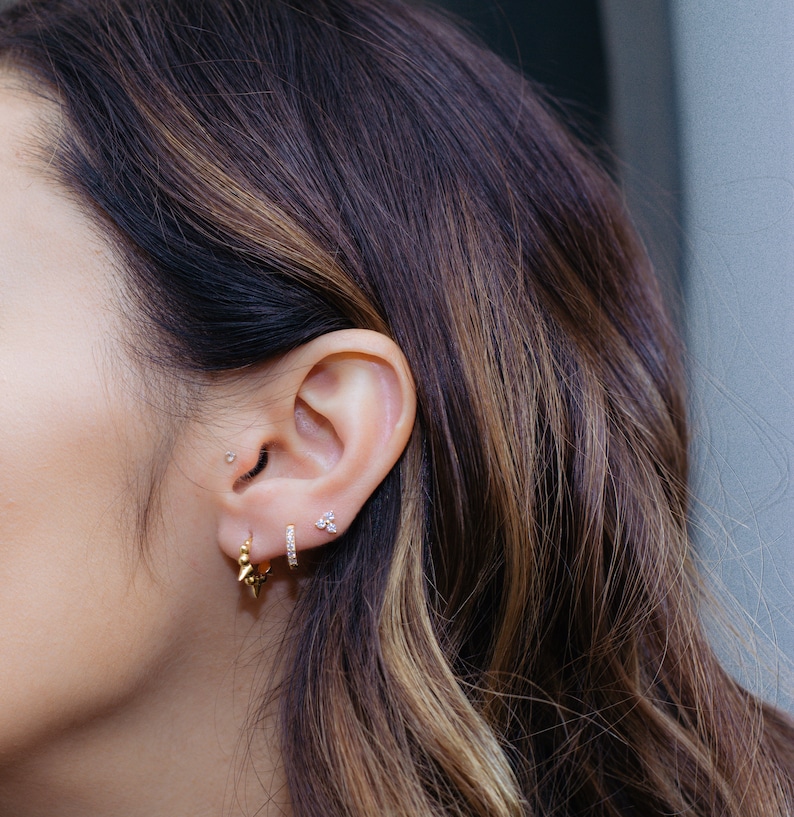 May include: A close-up of a person's ear with multiple earrings. The earrings are gold and have small diamonds. The person has brown hair.