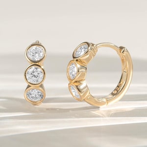 May include: Two gold hoop earrings, each with three clear, oval gemstones. The earrings are set against a light background, highlighting the polished gold and the sparkle of the gemstones. The design is simple and elegant, suitable for everyday wear.